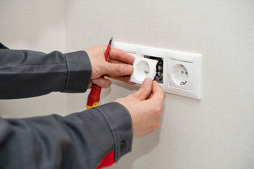 Electrician in uniform assembling electrical outlets on a white wall in a room.
